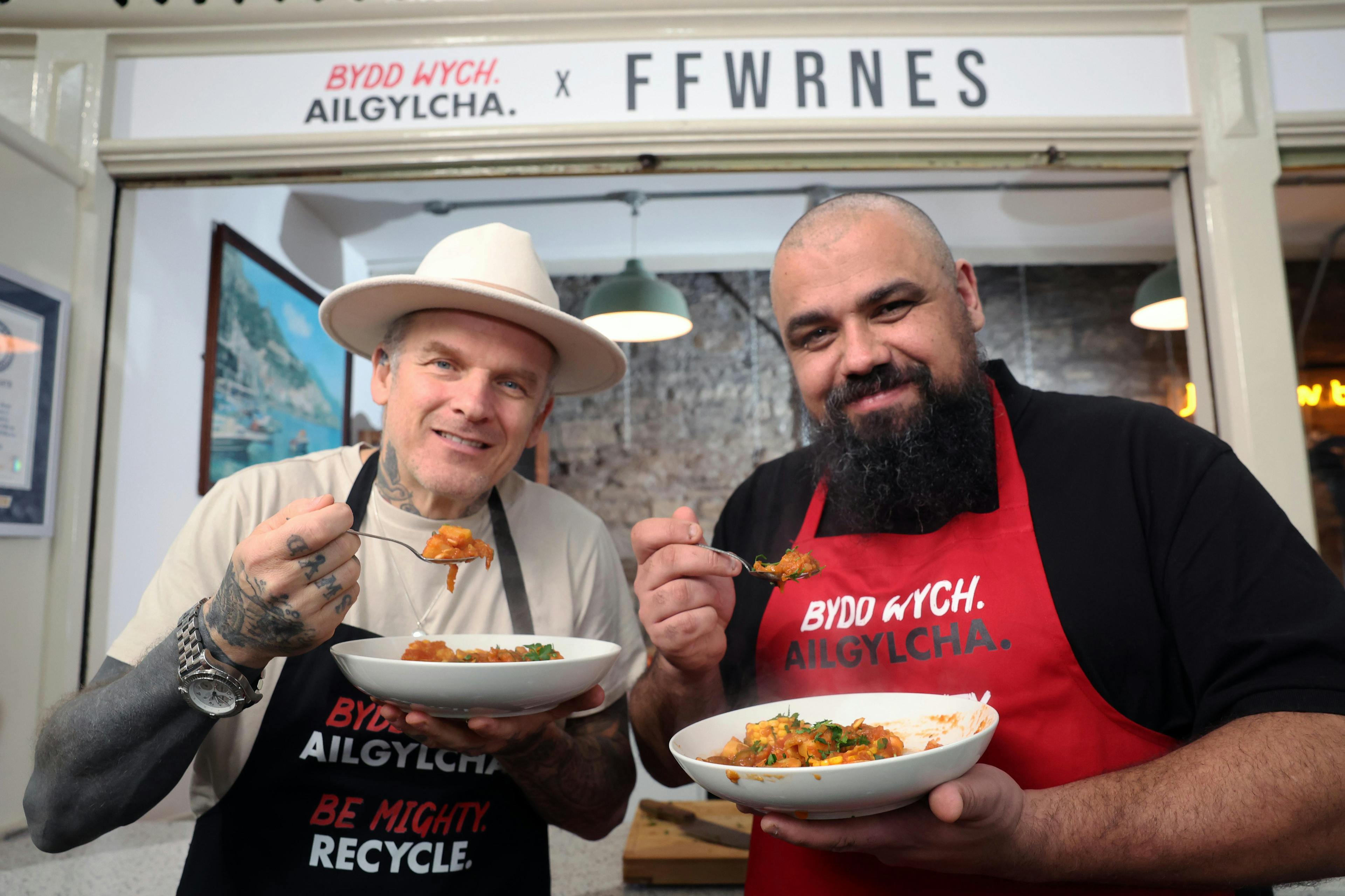 Two chefs standing in Cardiff Market holding white bowls of food and forks.