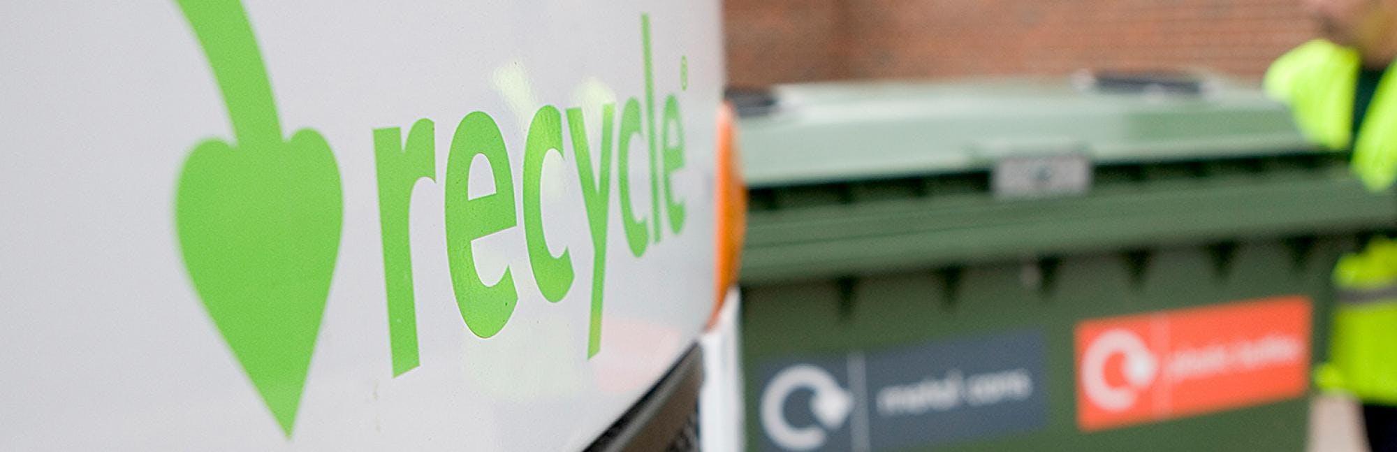 Close-up of a white vehicle with Recycle logo on the bonnet in green with recycling bins in the background