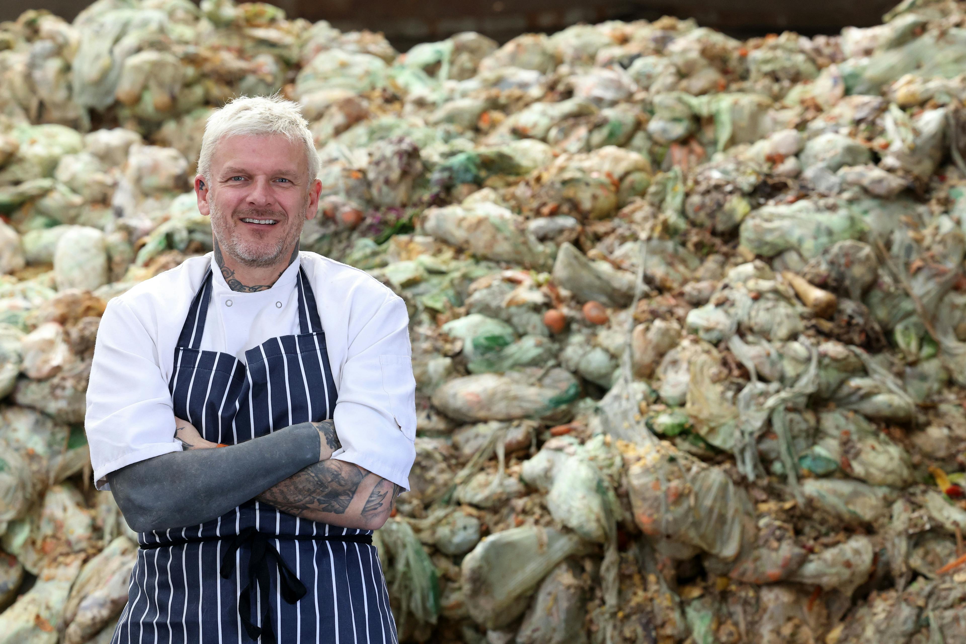 Matt Pritchard, a blonde white man with tattoos in a chef uniform, standing in from of a stack of full food recycling bags.